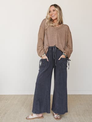 A smiling blonde model in a relaxed, lived-in outfit — oversized mocha v-neck sweater tucked loosely into wide-leg washed charcoal cargo pants with drawstring pockets, finished with neutral sandals and layered boho bracelets.