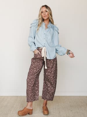 A light and earthy outfit pairing — a soft chambray ruffle-yoke button-down tucked loosely into wide-leg, spotted dark brown pants with a cream tie waist, finished with tan wooden clogs and stacked bracelets for that effortless, collected boho feel.