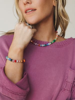 A close-up lifestyle shot of a multicolor beaded necklace and matching bracelet styled against a washed berry sweatshirt — the kind of jewelry that feels collected over time, not bought all at once.