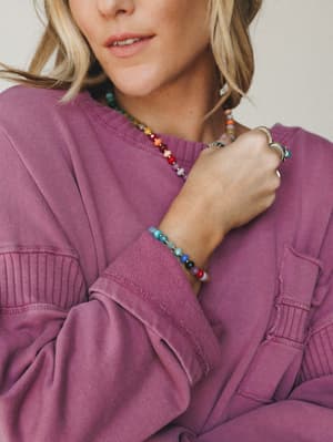 A close-up lifestyle shot of a model in a washed mauve sweatshirt, wearing a colorful multi-stone beaded necklace and matching bracelet stacked with a silver cuff — the kind of jewelry that looks like it has a story.