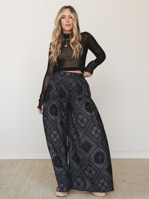 A blonde model in a studio setting wears a sheer black polka dot crop top with wide-leg bandana-print palazzo pants — dark, expressive, and effortlessly boho with layered gold jewelry and espadrille sandals.