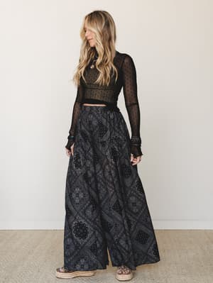A moody, bohemian full-body shot of a model in a sheer black dot-mesh crop top and a sweeping paisley-print maxi skirt, styled with layered gold jewelry and espadrille platform sandals — the kind of outfit that feels equally at home at a concert or a
