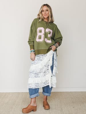 A relaxed, layered outfit built around unexpected contrasts — an olive varsity sweatshirt paired with a tiered lace midi skirt worn over cuffed jeans and wood-studded clogs, styled with stacked turquoise and pearl bracelets for that effortless boho-m