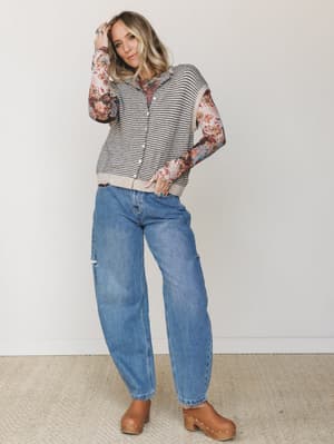 A relaxed, layered fall outfit — striped sweater vest over a floral long-sleeve top, styled with roomy distressed jeans and tan clogs, shot against a soft studio wall with an effortlessly lived-in feel.