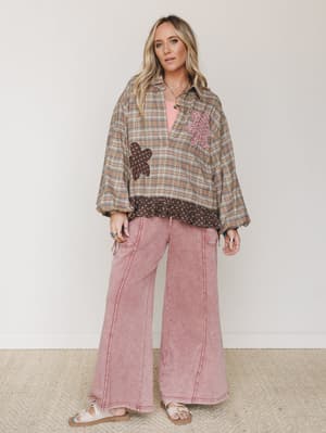A relaxed, layered outfit with a tan plaid oversized pullover shirt featuring floral appliqué patches, worn over a pink tank, paired with wide-leg acid wash rose pants and white sandals — the kind of put-together-but-not-trying look that feels genuin