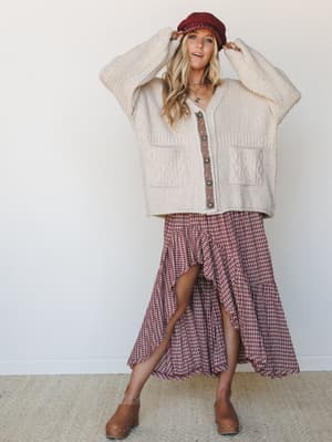 A breezy full-body shot of a model layering a chunky cream cable-knit cardigan over a plaid ruffle midi skirt, finished with a burgundy newsboy cap and tan clogs — the kind of autumn outfit that looks effortless but definitely took thought.