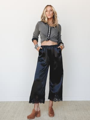 A knotted gray henley top paired with black satin wide-leg cropped pants trimmed in lace, styled with tan clogs and stacked bracelets — effortlessly cool with just enough edge.