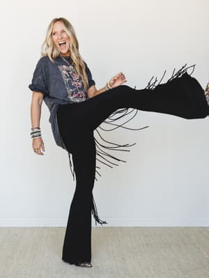 A laughing blonde woman kicks her leg up mid-air, showing off black fringe flare pants paired with a washed graphic tee — the kind of outfit that makes you feel like you should be at a concert you'll talk about for years.