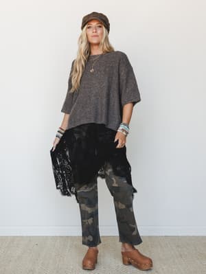 A full-body model shot with an effortlessly layered, unexpected mix — oversized knit sweater, black lace skirt layered over camo pants, and brown clogs — styled with a newsboy cap and stacked bracelets for a boho-meets-street vibe that feels personal
