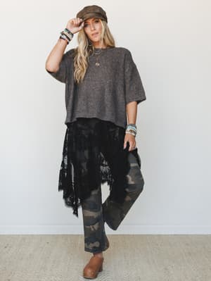 A full-body model shot of a layered boho-edgy look — oversized charcoal knit sweater, black lace skirt draped asymmetrically over camo flare pants, finished with a baker boy cap, stacked bracelets, and tan studded clogs.
