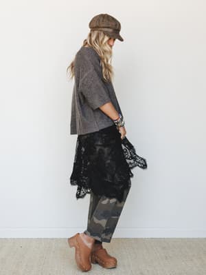A layered, textural look shot in profile — cropped knit sweater over a black lace midi skirt layered over camo pants, finished with a distressed newsboy cap and studded clogs, giving off that effortless 'I threw this together in 10 minutes and it som