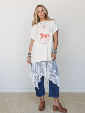 A layered boho outfit built around a cream graphic tee with a red horse print, worn over a flowy lace skirt and cropped dark denim jeans, styled with brown platform clogs and stacked beaded bracelets — free-spirited and effortlessly put-together.