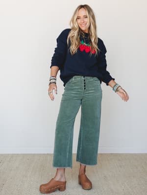 A smiling blonde model in a navy strawberry intarsia sweater and wide-leg sage green corduroy pants styled with tan studded clogs and stacked beaded bracelets — the kind of outfit that looks effortless but was absolutely thought about.