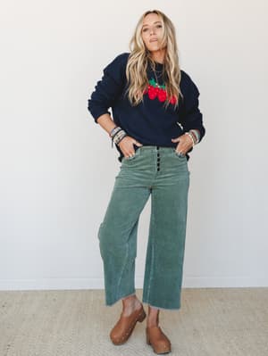 A relaxed, lived-in outfit with real personality — a navy strawberry intarsia sweater paired with sage wide-leg corduroy pants and tan clogs, styled with stacked beaded bracelets and layered gold necklaces for that effortless boho-casual feel.
