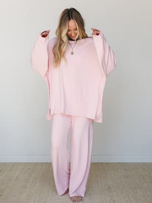 A matching two-piece set in the softest blush pink — oversized long-sleeve top with wide-leg pants worn barefoot against a clean studio wall, styled with layered necklaces and a smile that says she's not going anywhere today.