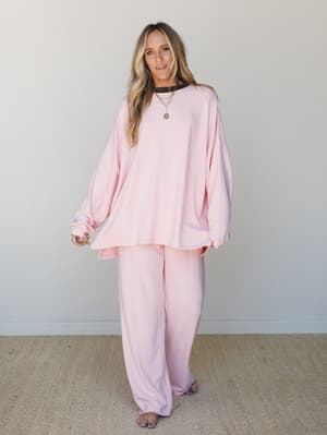 A matching oversized sweatshirt and wide-leg pants set in soft blush pink, styled barefoot with layered gold necklaces and a peeking chocolate brown tee underneath — the kind of outfit that makes staying home feel intentional.