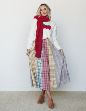 A cozy, collected fall outfit built around a patchwork plaid midi skirt in soft mixed plaids, layered with a cream strawberry intarsia sweater and a chunky red knit scarf — the kind of outfit that feels like it was put together intuitively and someho