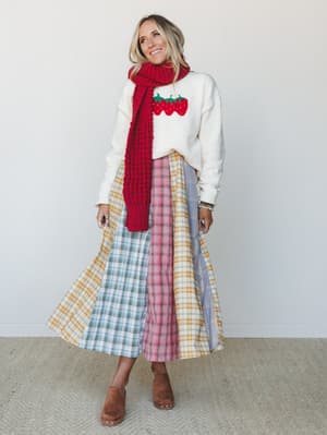 A smiling blonde model layers a cream strawberry graphic sweatshirt with a chunky red knit scarf and a full patchwork plaid midi skirt in mixed plaids, finished with tan suede clogs — the kind of outfit that looks like it came together effortlessly b