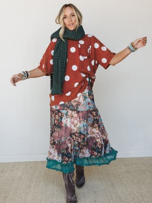 A full-body model shot with a playful, mixed-pattern energy — oversized rust polka dot tee layered over a patchwork floral midi skirt with a teal ruffle hem, finished with a chunky knit scarf and dark cowboy boots.
