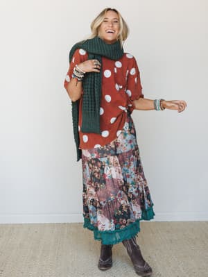 A laughing model styled in a mix-and-match boho bundle — polka dot oversized top, patchwork floral maxi skirt with a teal ruffle underlayer, and a chunky knit scarf — shot against a clean studio wall with an easy, joyful energy.