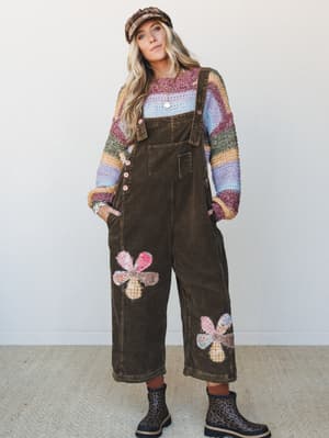 A full-body model shot of a layered boho outfit built around wide-leg corduroy overalls in deep brown, styled with a multicolor striped knit sweater, a plaid newsboy cap, and leopard print rain boots — the patchwork flower appliqués on the overalls d