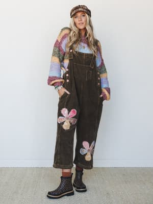 A layered boho look built around wide-leg corduroy overalls in deep brown, styled with a rainbow striped knit sweater, plaid newsboy cap, and leopard print boots — the kind of outfit that looks like it came together effortlessly but was absolutely th