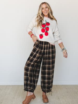 A smiling blonde woman in a cream tomato-graphic sweater and dark plaid jogger pants styled with tan clogs — the kind of outfit that makes a Saturday errand run feel like a personality statement.
