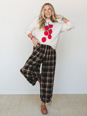 A smiling model in a cream novelty knit sweater with a tomato intarsia graphic paired with relaxed plaid jogger pants and tan mules — the kind of outfit that feels like autumn in the best possible way.