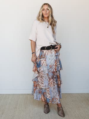 A relaxed, put-together boho look built around a flowy tiered midi skirt in a blue and rust floral patchwork print, tucked under with an oversized cream tee cinched by a studded black belt — finished with leopard mules and layered jewelry for that ef