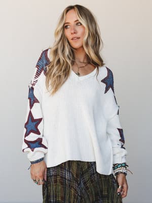 A relaxed, expressive look anchored by an oversized cream knit sweater with bold star-print sleeves, layered over an olive plaid skirt and finished with stacked turquoise and beaded bracelets — the kind of outfit that feels effortless but clearly tho