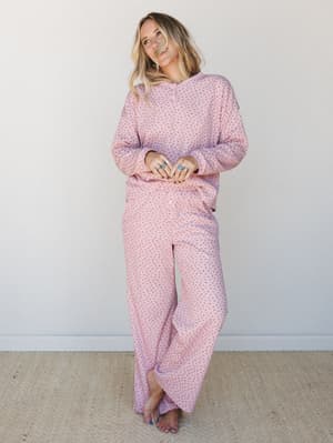 A matching pink floral two-piece set — long-sleeve top and wide-leg pants — worn barefoot against a soft white wall, giving off total cozy Sunday morning energy.