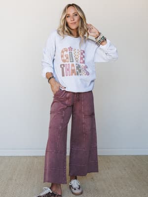 A relaxed, feel-good fall outfit — an oversized 'Give Thanks' graphic sweatshirt paired with wide-leg plum mineral-wash pants, styled with leopard sneakers and a stack of bracelets for that effortlessly put-together everyday look.