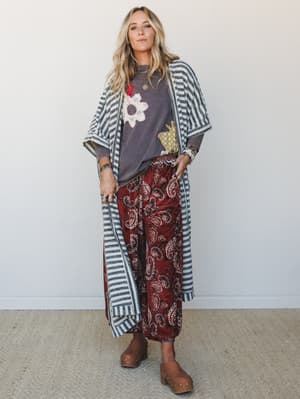 A layered boho outfit built on texture and pattern-mixing — striped duster cardigan over a graphic tee with floral and frog appliqué, paired with wide-leg paisley pants and wooden clogs, shot against a clean studio wall.