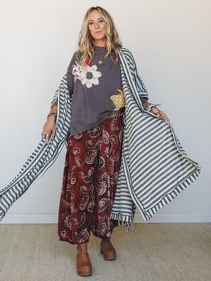 A layered, mix-pattern look full of personality — floral appliqué graphic tee, wide-leg paisley pants in deep burgundy, and a dramatic striped blanket wrap held open like a cape, grounded with chunky platform clogs.