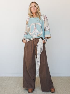 A relaxed full-body shot of a model in a patchwork thermal peace sign sweatshirt and wide-leg chocolate brown pants, styled with a lace sash at the waist and stacked bracelets — the kind of outfit that looks effortless but clearly wasn't an accident.