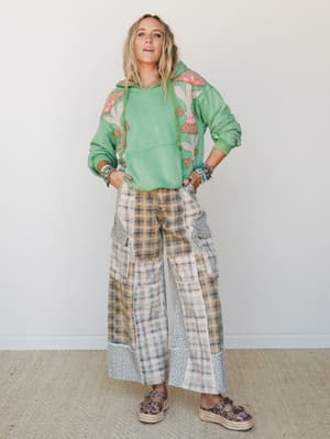 A full-body studio shot of a model in a green mushroom-embroidered hoodie and wide-leg patchwork plaid cargo pants, styled with platform sandals and stacked bracelets — the kind of outfit that feels effortlessly weird in the best way.