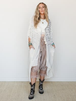 A flowy ivory sequin-embellished duster kimono styled over a cropped white top and dusty blush pleated pants with lace trim, finished with embroidered black western boots and stacked turquoise bracelets — the kind of outfit that feels like it has a s