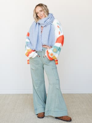 A relaxed, color-happy outfit layered with a multicolor granny square cardigan, a soft periwinkle fringe scarf, and wide-leg sage flare pants — the kind of put-together-but-cozy look that feels like a good mood in clothing form.