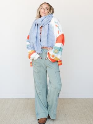 A relaxed full-body studio shot of a model layered in a colorful granny square and striped knit cardigan, a periwinkle fringe scarf, and wide-leg sage green pants with seam detailing — the kind of outfit that feels like a warm Saturday with nowhere t