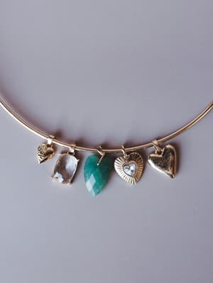 A gold wire collar necklace hung with five mixed charms — a textured leaf, a clear faceted stone, a teal heart-shaped gemstone, a sunburst crystal heart, and a smooth gold heart — shot flat against a muted lavender-gray background with a collected, s