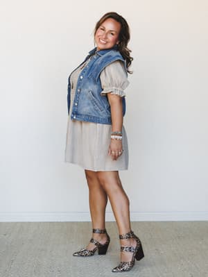 A smiling model poses in profile wearing a breezy sand-colored mini dress layered under a cropped denim vest, finished with leopard print block-heel sandals and a stack of beaded bracelets — the kind of outfit that looks effortless but definitely too