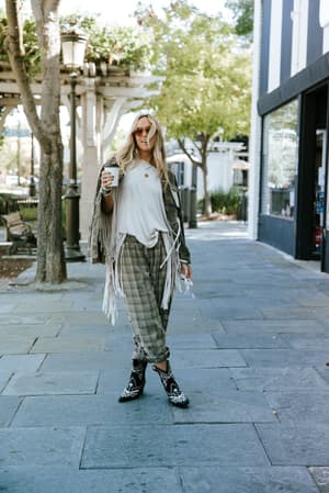 A sun-lit sidewalk moment — she's holding her coffee, wearing plaid relaxed pants, a white tee, and a fringe jacket draped over her shoulders, grounded by embroidered western boots that do all the talking.