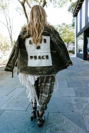 Shot from behind on a sun-dappled sidewalk, this look layers a camo fringe jacket with a 'Love & Peace' patch over plaid joggers and embroidered western boots — the kind of outfit that turns a coffee run into a whole moment.