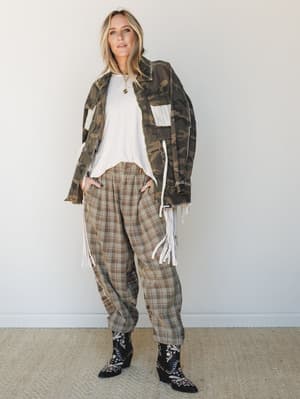 A relaxed, lived-in layered look — camo jacket with fringe detail draped over a white tee, paired with plaid wide-leg trousers and embroidered western boots, shot against a clean studio wall with an effortlessly cool, free-spirited energy.