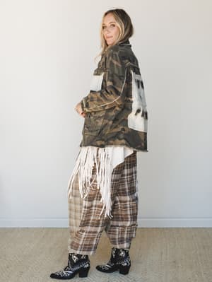 A full-body side profile of a model in a layered, texture-mixed outfit — camo utility jacket with patchwork and fringe details over a fringe top and oversized plaid pants, finished with black embroidered western boots.