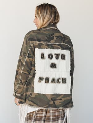 Shot from behind, a model wears an oversized camo utility jacket with a raw-edge cream patch reading 'Love & Peace' on the back, layered over a fringe top and plaid skirt — the kind of jacket that does all the talking.