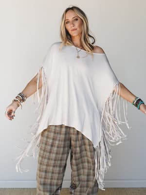 A relaxed, free-spirited look featuring an oversized white fringe poncho layered over wide-leg plaid pants, styled with stacked bracelets and a pendant necklace — effortless boho energy shot against a clean studio wall.