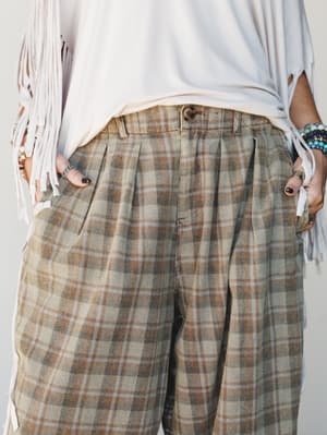 A close-up mid-shot of wide-leg plaid trousers in muted sage and warm brown tones, styled with a white fringe top and layered bracelets — the kind of outfit that feels effortlessly pulled-together without trying too hard.