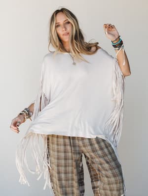 A flowy oversized fringe top paired with plaid wide-leg pants and stacked beaded bracelets — the kind of outfit that looks effortless but totally intentional, shot against a soft neutral studio backdrop.