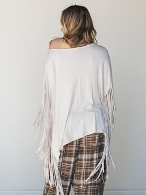 Shot from behind, a model wears a cream fringe poncho layered over a mustard bralette and brown plaid wide-leg pants — the kind of effortless, thrown-together outfit that somehow looks completely intentional.